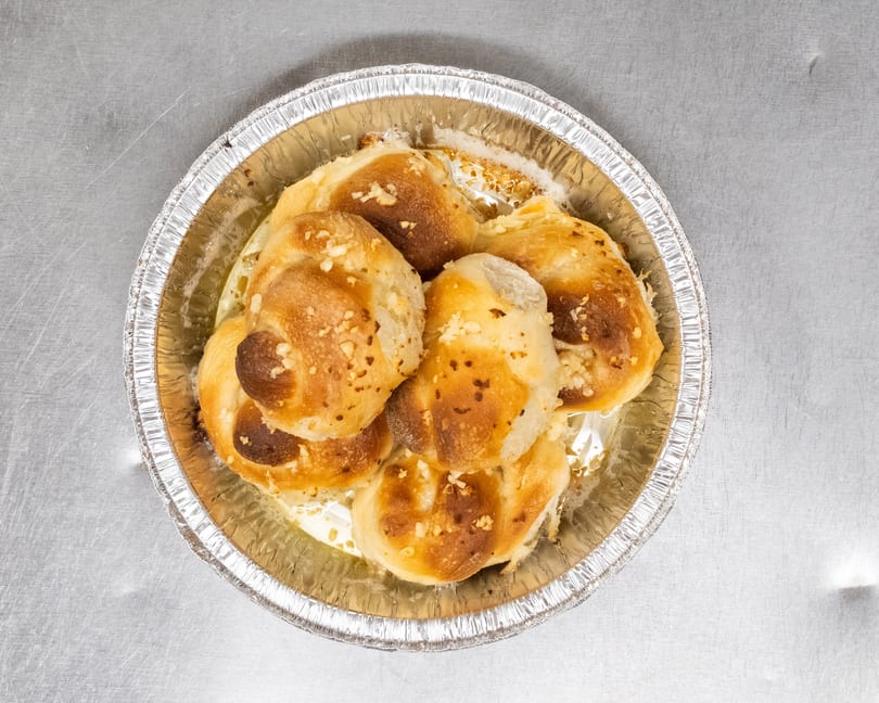 Golden-brown garlic knots, glistening with butter and sprinkled with garlic, in a foil pan.