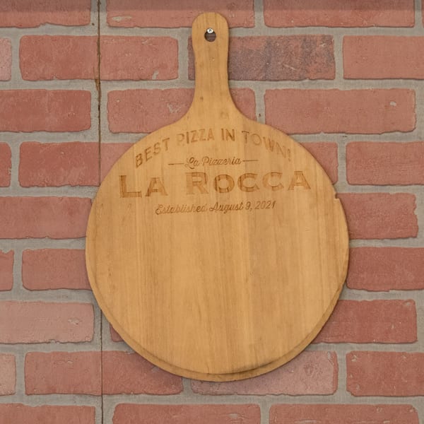 Wooden pizza peel with "Best Pizza in Town! La Pizzeria La Rocca" engraved, hanging on a brick wall.