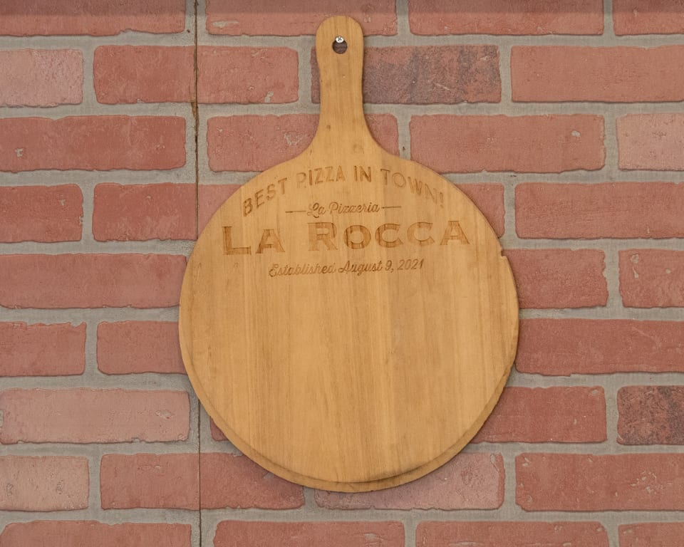 Wooden pizza peel with "Best Pizza in Town! La Pizzeria La Rocca" engraved, hanging on a brick wall.