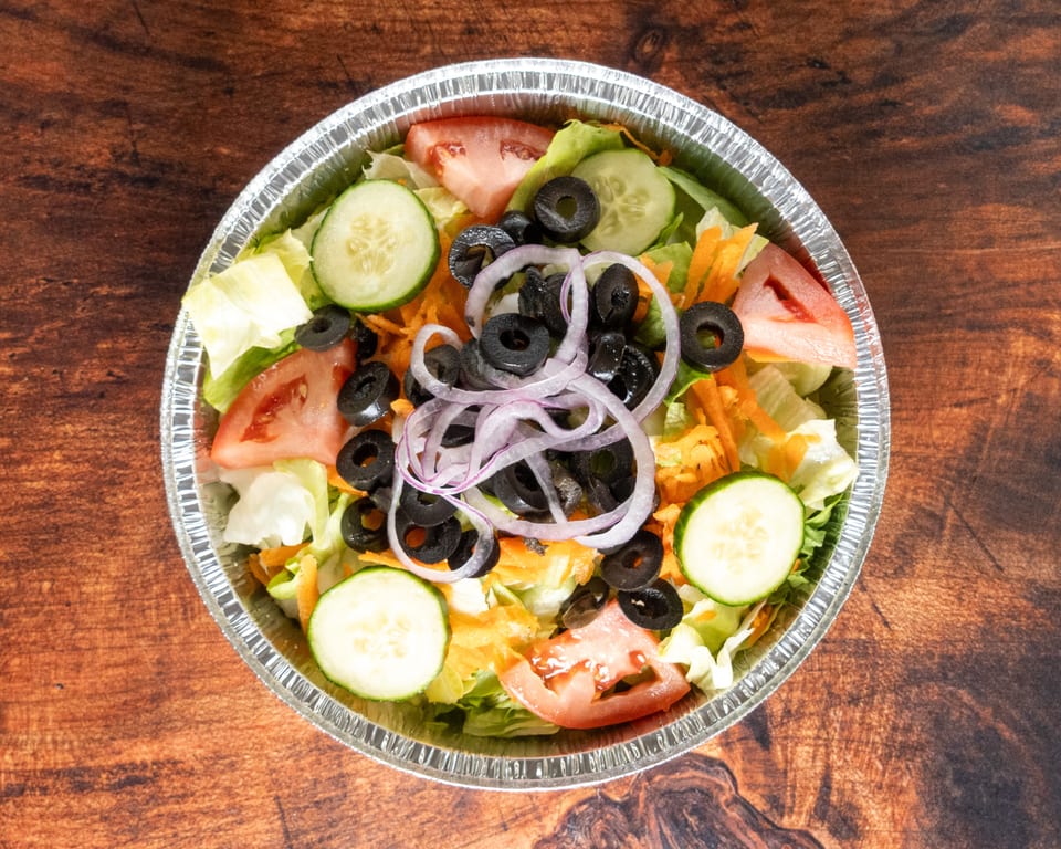 Fresh garden salad with lettuce, tomatoes, cucumbers, black olives, and red onion in a foil container.