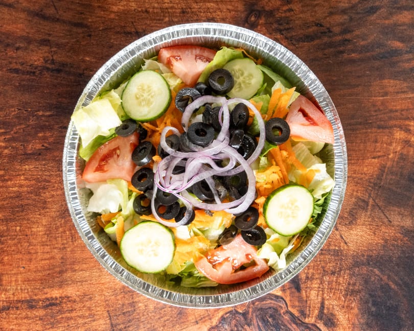 Fresh garden salad with lettuce, tomatoes, cucumbers, black olives, and red onions in a foil container.