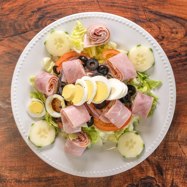 Chef salad with ham, salami, hard-boiled eggs, olives, tomatoes, and cucumbers on a white plate.