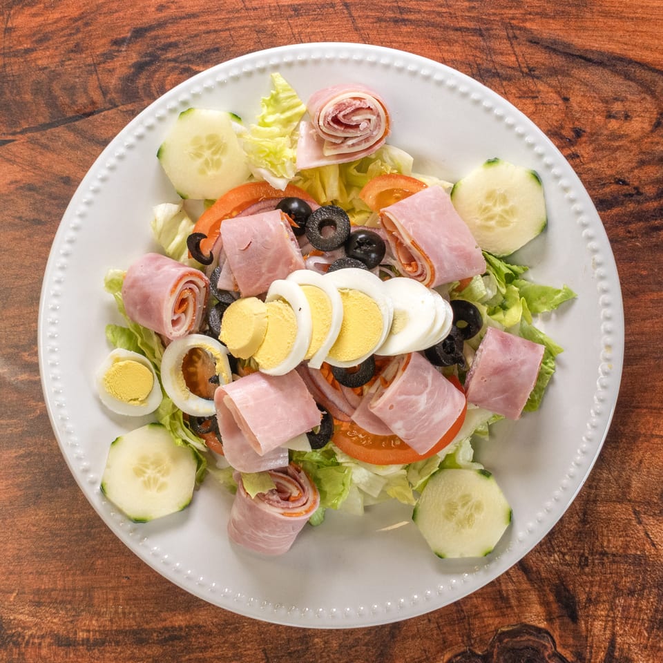 Chef salad with ham, salami, hard-boiled eggs, olives, tomatoes, and cucumbers on a white plate.
