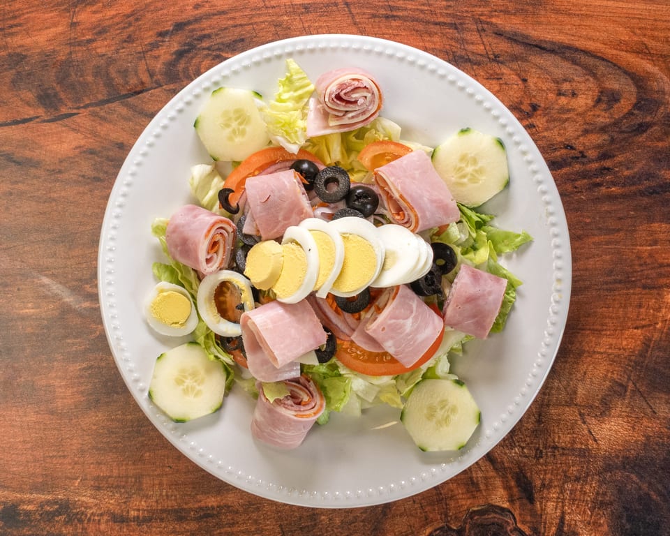 Chef salad with ham, salami, hard-boiled eggs, olives, tomatoes, and cucumbers on a white plate.