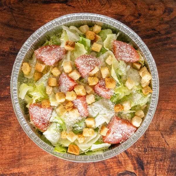 Caesar salad with lettuce, tomatoes, croutons, and parmesan cheese in a foil container.
