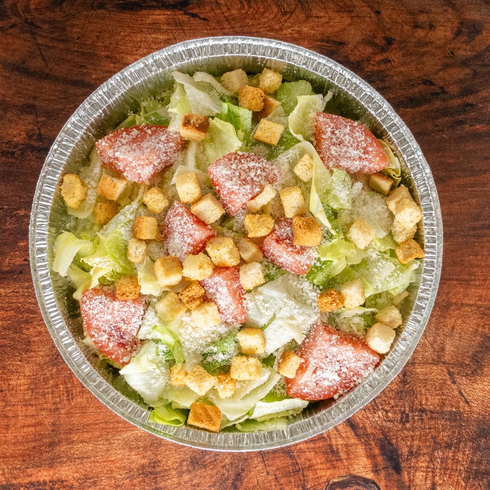 Caesar salad with lettuce, tomatoes, croutons, and parmesan cheese in a foil container.
