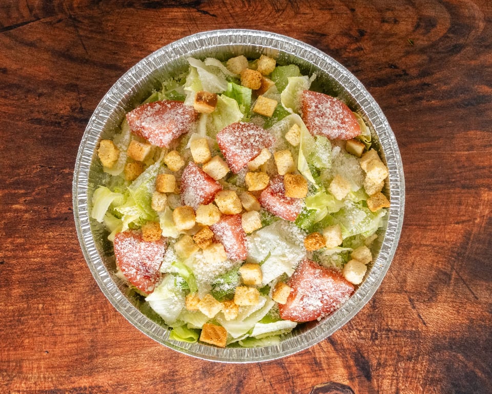 Caesar salad with lettuce, tomatoes, croutons, and parmesan cheese in a foil container.