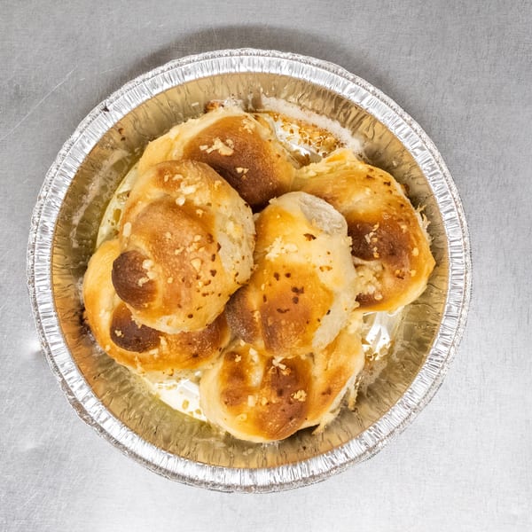 Golden-brown garlic knots, glistening with butter and sprinkled with garlic, in a foil pan.
