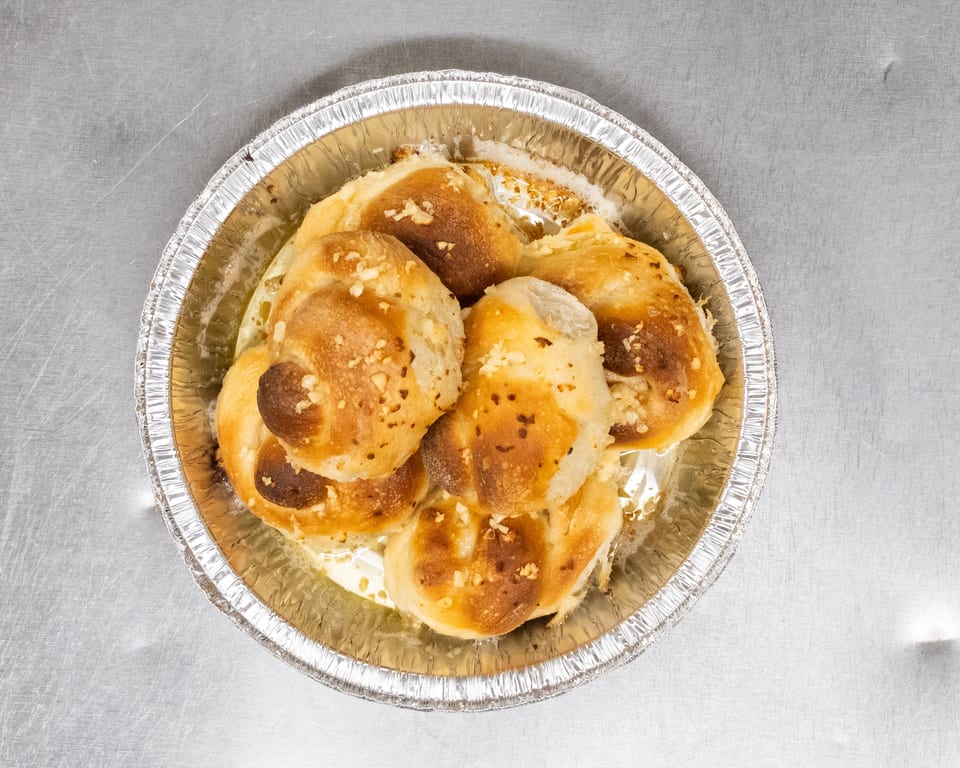 Golden-brown garlic knots, glistening with butter and sprinkled with garlic, in a foil pan.