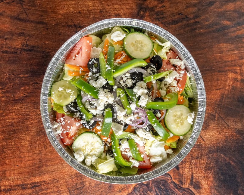 A vibrant salad with lettuce, tomatoes, cucumbers, olives, green peppers, and feta cheese in a foil container.