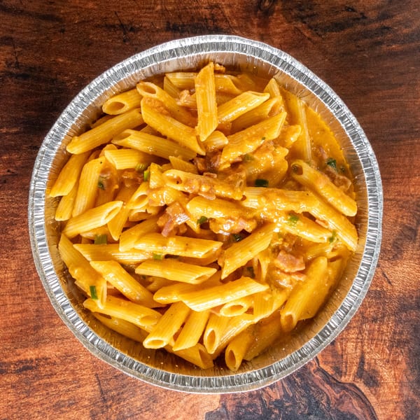 Penne pasta with creamy orange sauce, bacon, and green onions in a foil container.