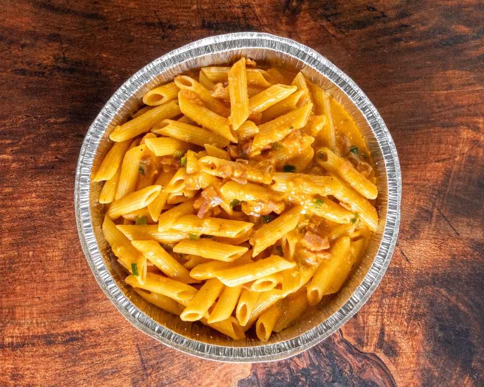 Penne pasta with creamy orange sauce, bacon, and green onions in a foil container.