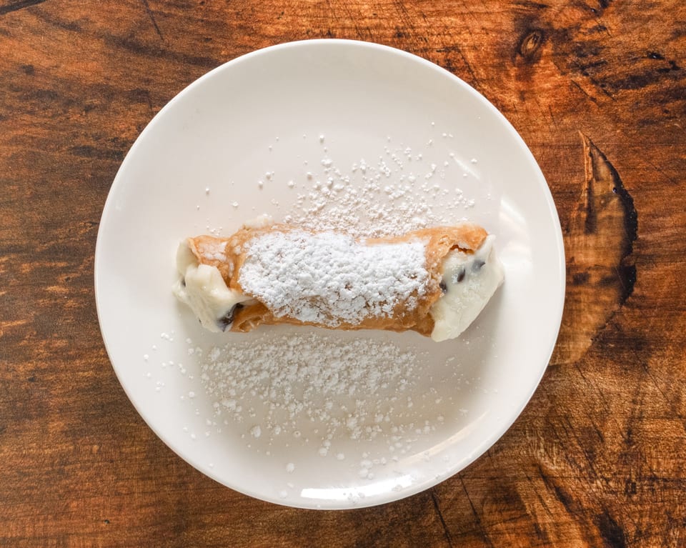 A single cannoli pastry, filled with cream and chocolate chips, dusted with powdered sugar on a white plate.