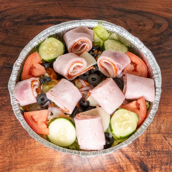 Antipasto salad in a foil container with ham and cheese rolls, tomatoes, cucumbers, and olives.